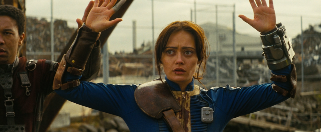 Ella Purnell as Lucy MacLean and Aaron Moten as Maximus on Fallout, shown crossing a bridge slowly with their arms raised in the air.